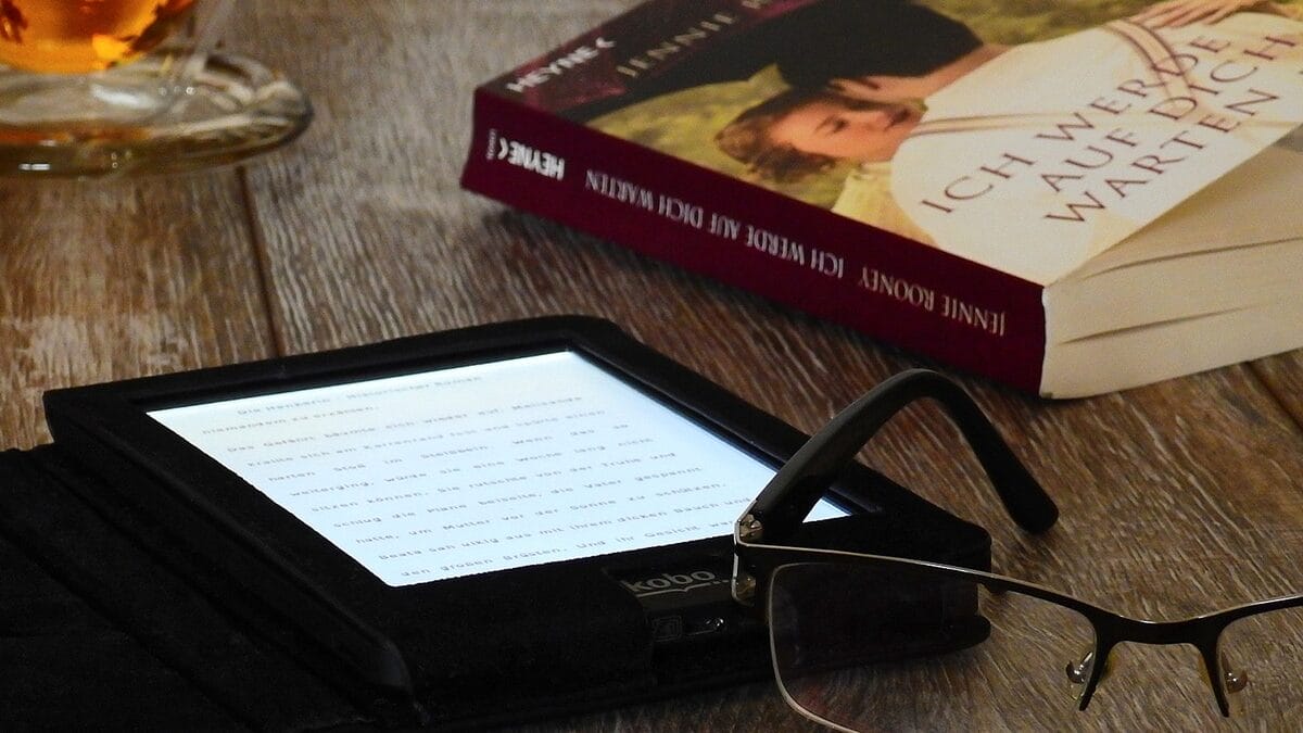 Un livre à couverture rigide, une liseuse, des lunettes et une tasse de thé disposés sur une surface en bois.