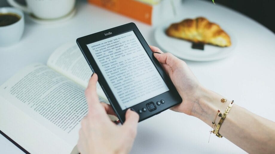 Une personne tenant une liseuse Kindle à côté d'un livre physique et une tasse de café avec un croissant sur une table blanche.