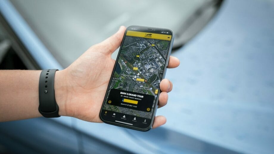 Une main tenant un smartphone affichant une carte avec des emplacements GPS sur l'écran, suggérant une navigation ou une application de guide touristique.  L’arrière-plan présente une vue floue d’un tableau de bord de voiture.
