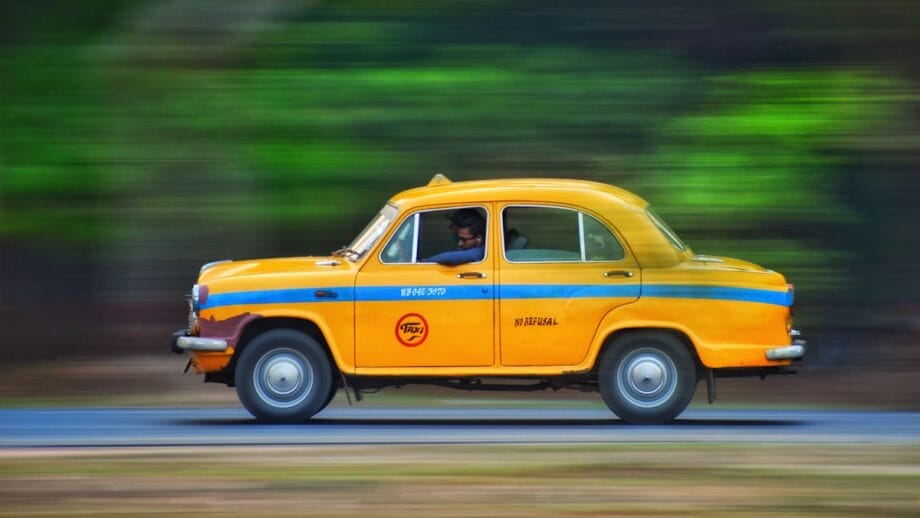 Un taxi jaune avec des rayures noires et le mot « taxi » imprimé dessus est en mouvement dans une rue floue, indiquant une technique de photographie panoramique pour capturer la sensation de vitesse.