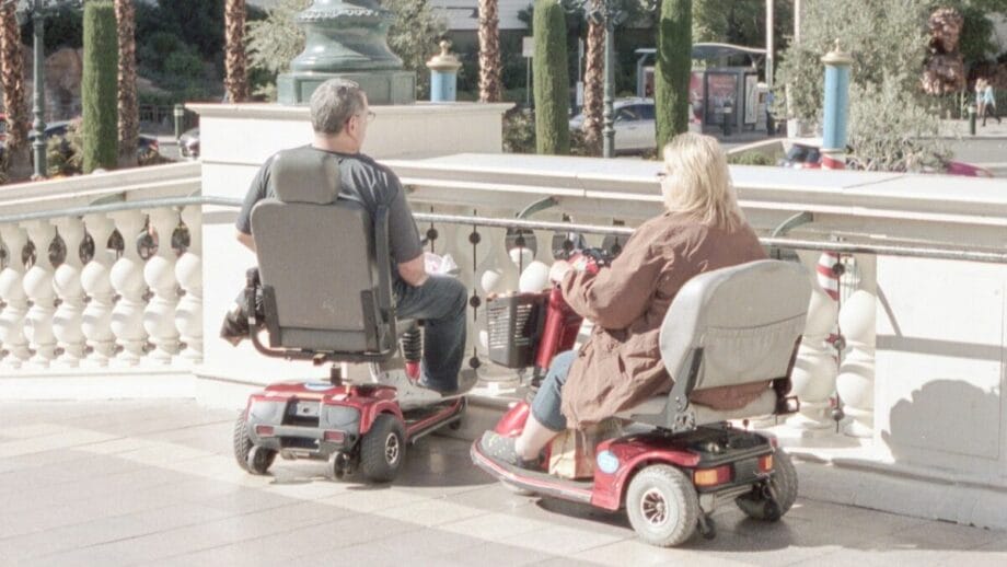 Deux personnes sur des scooters de mobilité sont arrêtées sur une zone pavée à côté d'une balustrade décorative avec des palmiers et des bâtiments en arrière-plan.