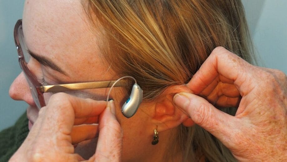 Une personne portant des lunettes se fait poser une aide auditive derrière l'oreille par les mains d'une autre personne.