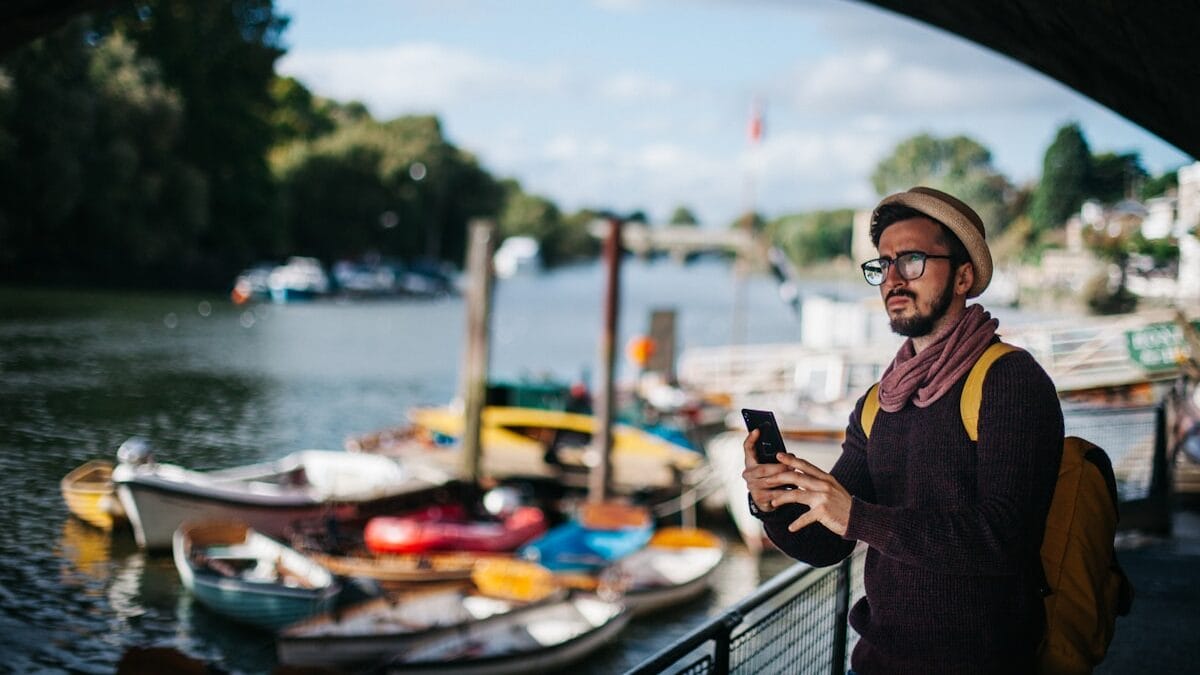Un homme avec un chapeau et des lunettes se tient sous un pont au bord d'une rivière, regardant son téléphone. Plusieurs bateaux sont amarrés à proximité sous un ciel partiellement nuageux.