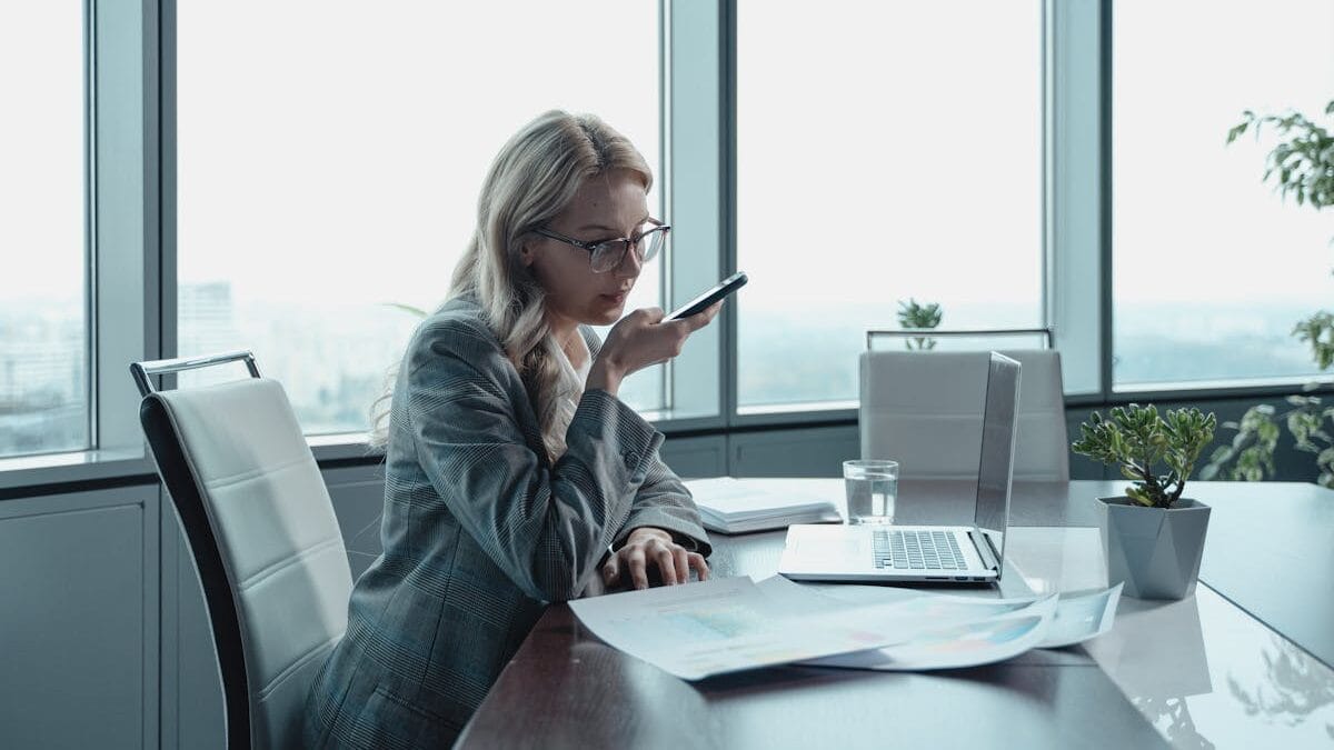 Une femme en costume d'affaires parle dans son téléphone à une table de conférence avec un ordinateur portable et des documents devant elle, dans un bureau lumineux.