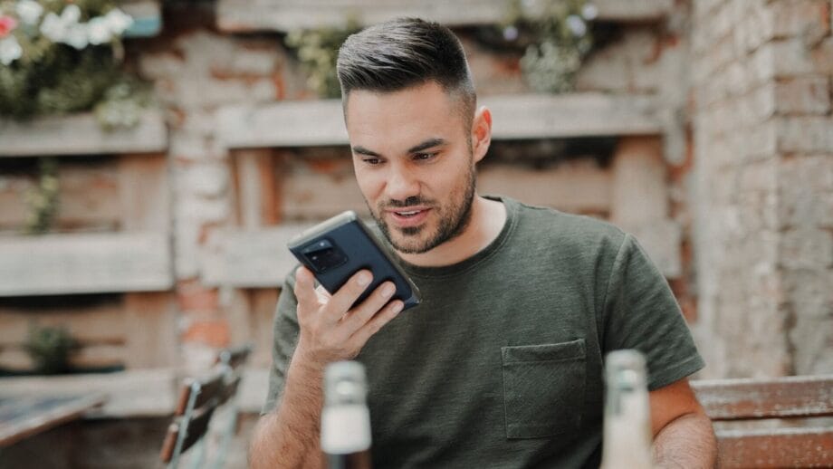 Un homme regarde son téléphone et parle à l’aide du haut-parleur alors qu’il est assis à une table.