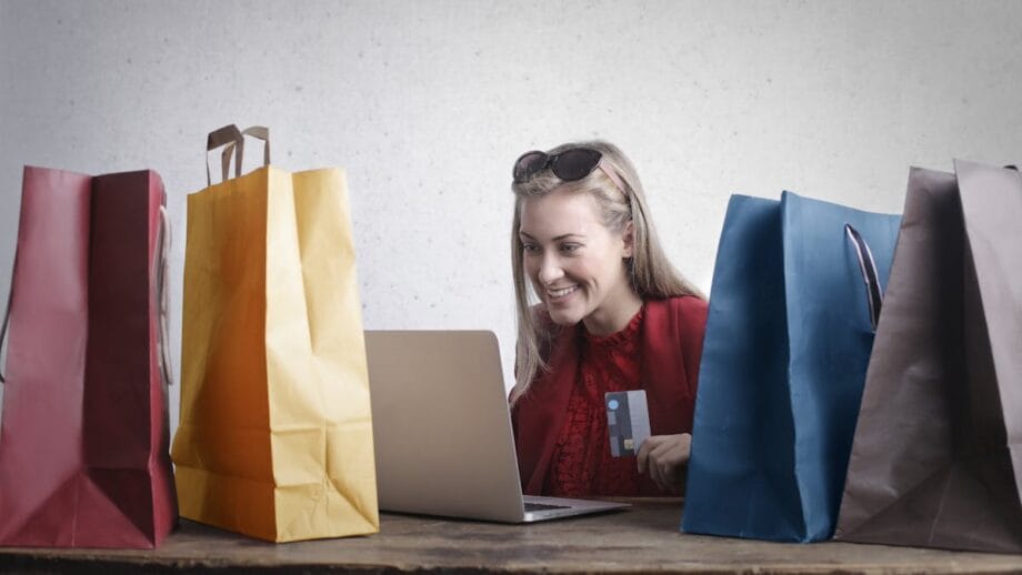 Une femme est assise à une table avec plusieurs sacs à provisions, utilisant un ordinateur portable et tient une carte de crédit.