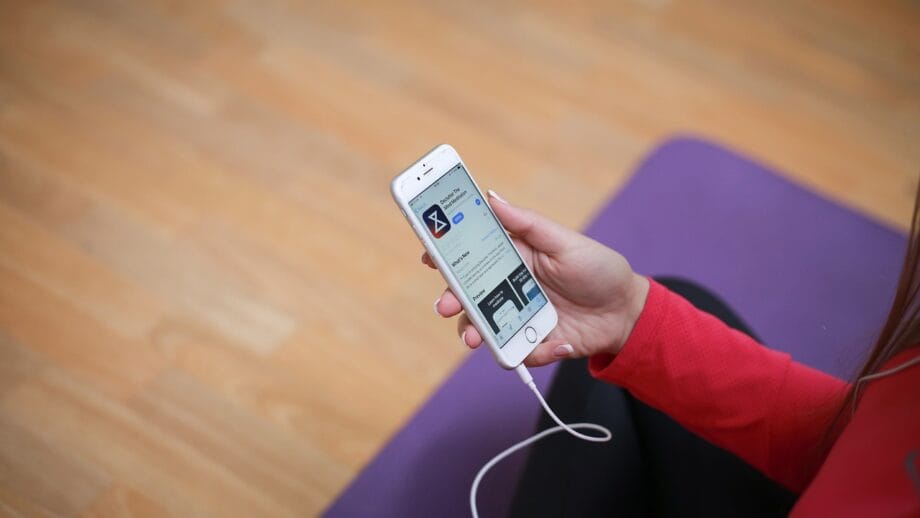Personne tenant un smartphone affichant une application, avec des écouteurs branchés. Ils sont assis sur un coussin violet sur un plancher en bois.