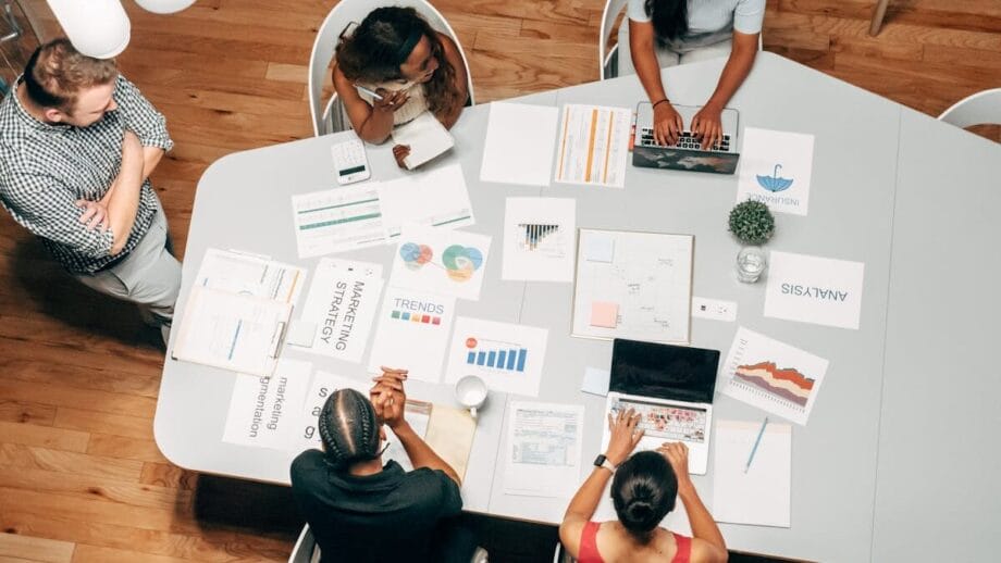 Un groupe de cinq personnes travaillant à une table avec des ordinateurs portables, des graphiques et des articles, engageant une discussion ou une réunion.