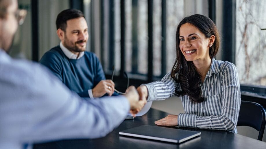 Deux professionnels servent la main sur une table, concluant probablement une réunion ou un accord, tandis qu'un troisième individu observe l'interaction avec un sourire.
