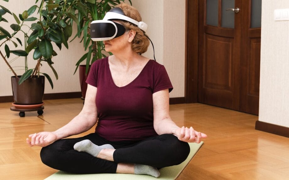 Une femme est assis en tailleur croisée sur un tapis de yoga portant des lunettes VR. Elle a les mains reposant sur ses genoux, entourées de plantes intérieures dans une pièce avec un parquet en bois.
