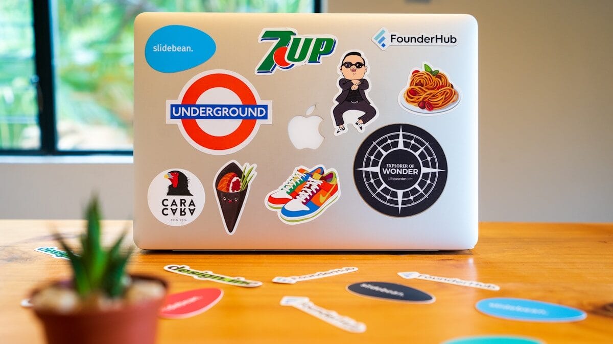 Ordinateur portable recouvert de divers autocollants colorés, dont 7up, underground et autres, sur une table en bois avec des autocollants dispersés à proximité; Une petite plante est au premier plan.