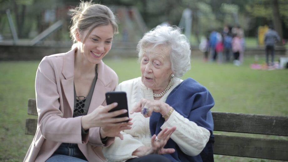 Deux femmes assises sur un banc de parc, l'une pointant sur un écran de smartphone tandis que l'autre sourit.