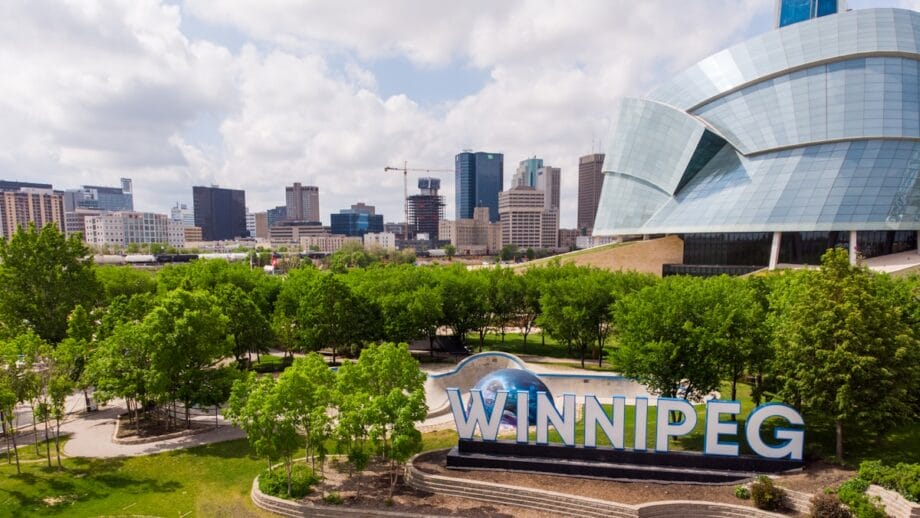 Horizon de la ville de Winnipeg avec des bâtiments modernes, des arbres verts et un grand signe «Winnipeg» au premier plan un jour partiellement nuageux.