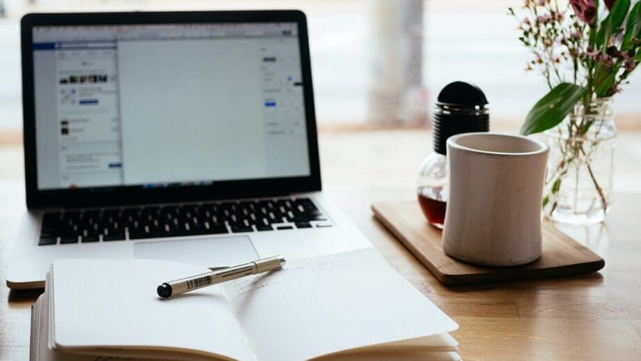 Open ordinateur portable, cahier avec stylo, tasse à café et fleurs sur un bureau en bois, suggérant un espace de travail ou une zone d'étude.