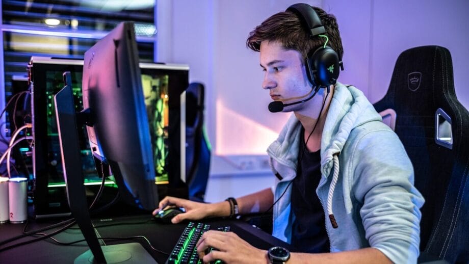 Un jeune homme portant des écouteurs et un microphone se trouve sur un bureau, jouant sur un ordinateur avec un clavier rétro-éclairé et un grand moniteur.