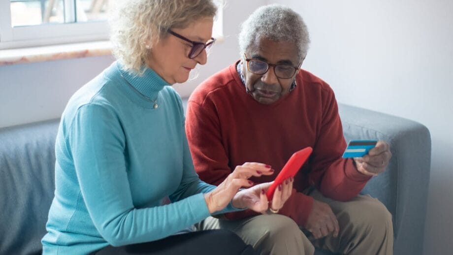 Deux adultes plus âgés sont assis sur un canapé; L'un détient un smartphone et l'autre détient une carte de crédit, semblant effectuer une transaction en ligne.