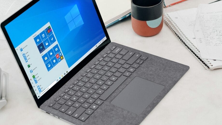 Un ordinateur portable avec un clavier gris et un écran de démarrage Windows 10 affiché. A proximité se trouvent un cahier avec des notes manuscrites, un cahier fermé et une tasse colorée.