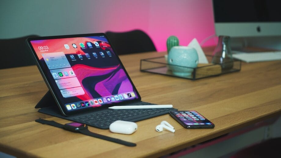 Un espace de travail moderne comprenant un iPad avec un clavier sur un support, un iPhone, un Apple Pencil, des Airpods et un ordinateur de bureau en arrière-plan, le tout soigneusement disposé sur un bureau avec un éclairage ambiant.