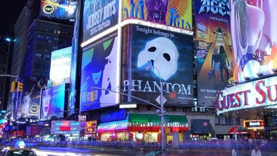Times Square la nuit, avec des panneaux d'affichage et des publicités lumineux, dont un pour "Phantom". Des traînées lumineuses provenant de voitures en mouvement sont visibles dans la rue en contrebas.