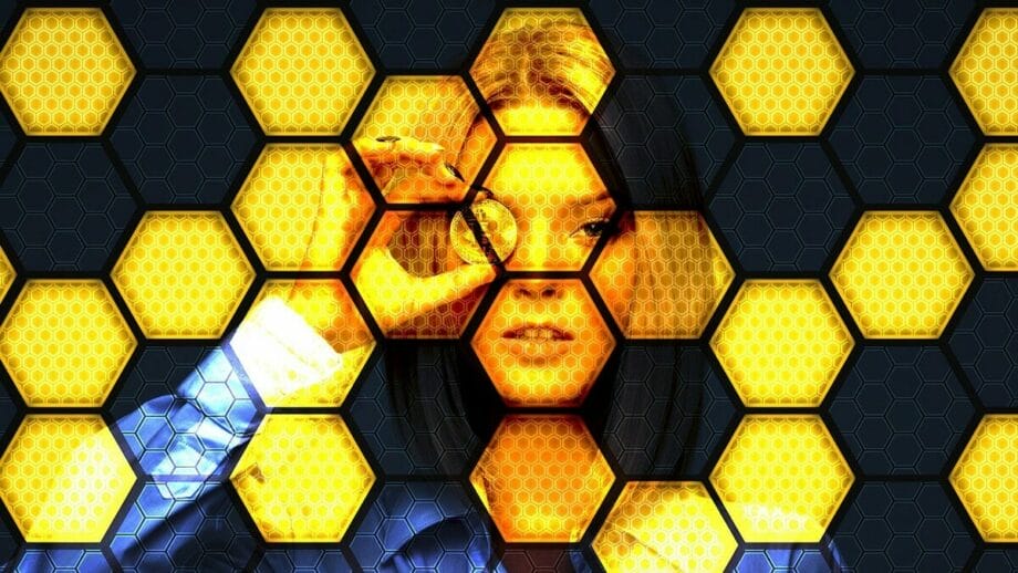 Une femme tient une pièce de monnaie près de son œil, vue à travers un motif en nid d'abeille avec des hexagones jaunes et noirs.