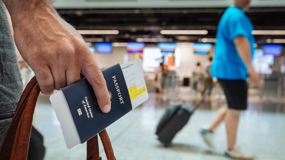 Une personne détient un passeport américain et une carte d'embarquement dans un terminal d'aéroport avec des voyageurs et des comptoirs d'enregistrement en arrière-plan.