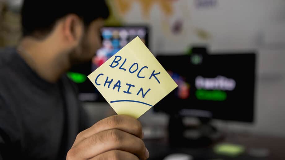 Une personne tient un post-it jaune sur lequel est écrit BLOCK CHAIN, avec des écrans d'ordinateur visibles en arrière-plan.