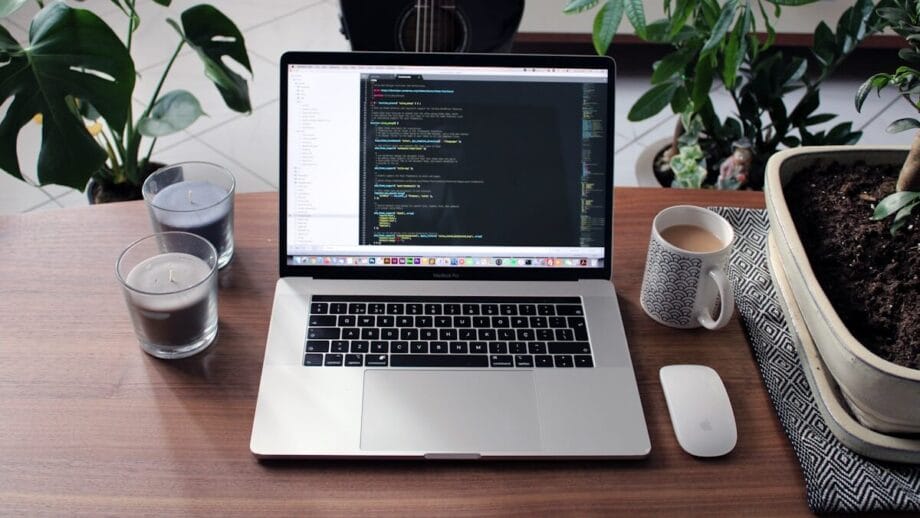 Un ordinateur portable affichant un code est posé sur une table en bois avec des bougies, une tasse, une souris d'ordinateur, des plantes d'intérieur et une guitare en arrière-plan.