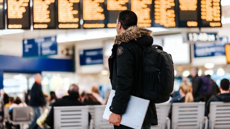 Une personne vêtue d'une veste et d'un sac à dos regarde un panneau de départ à l'aéroport, pensant à un voyage d'affaires réussi tout en tenant un ordinateur portable. D'autres personnes sont assises à l'arrière-plan.