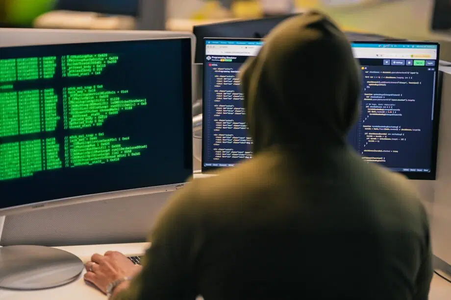 Une personne en sweat à capuche est assise à un bureau avec deux moniteurs affichant du code et du texte vert sur fond sombre.