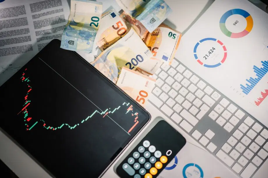 Un clavier d'ordinateur, une tablette avec un graphique boursier, une calculatrice, des billets en euros et des rapports financiers avec des tableaux et des graphiques.