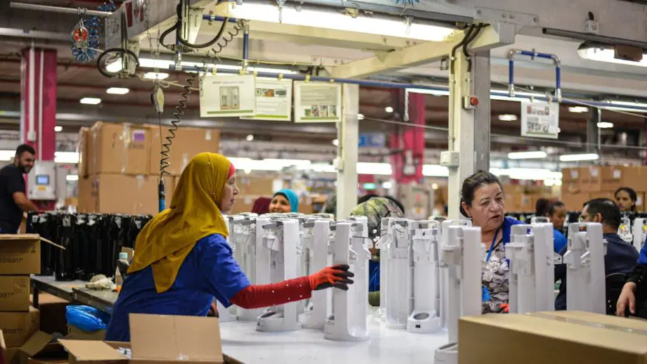 Les ouvriers de l'usine assemblent des appareils électroménagers blancs sur un établi, avec des boîtes et des machines visibles en arrière-plan.