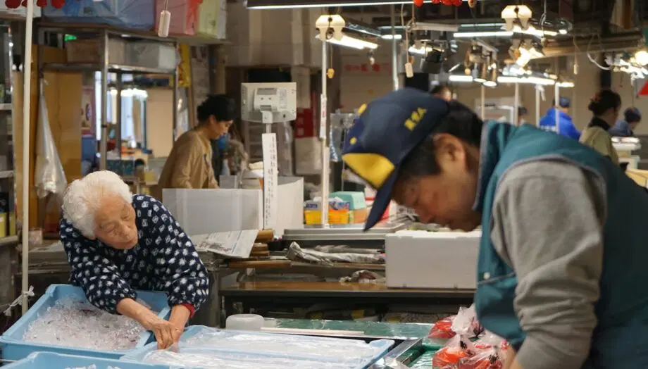 Une femme âgée range les articles dans un bac rempli de glace pendant qu'un travailleur manipule des marchandises dans un marché couvert très fréquenté.