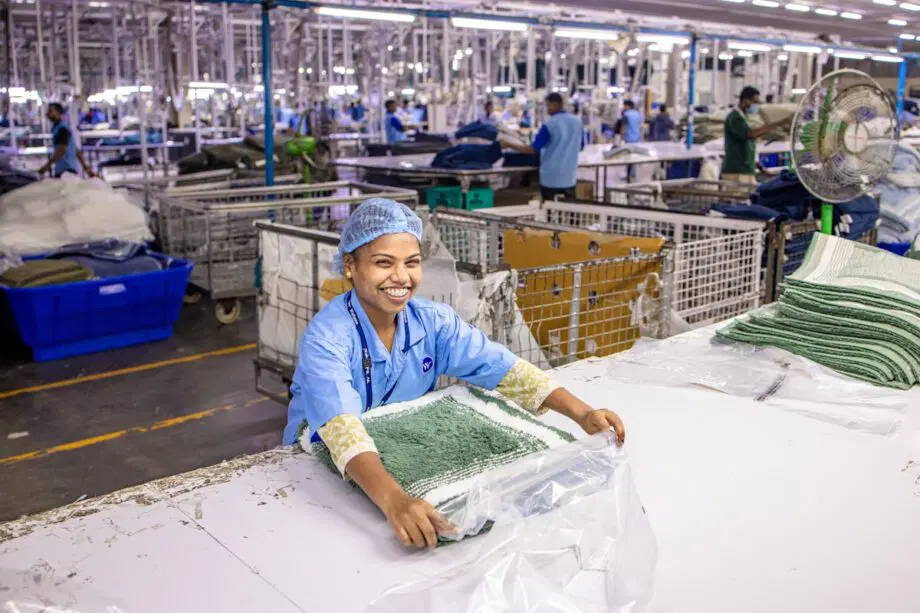 Un ouvrier en uniforme bleu et en filet à cheveux plie les serviettes à une table dans une usine textile bien éclairée.