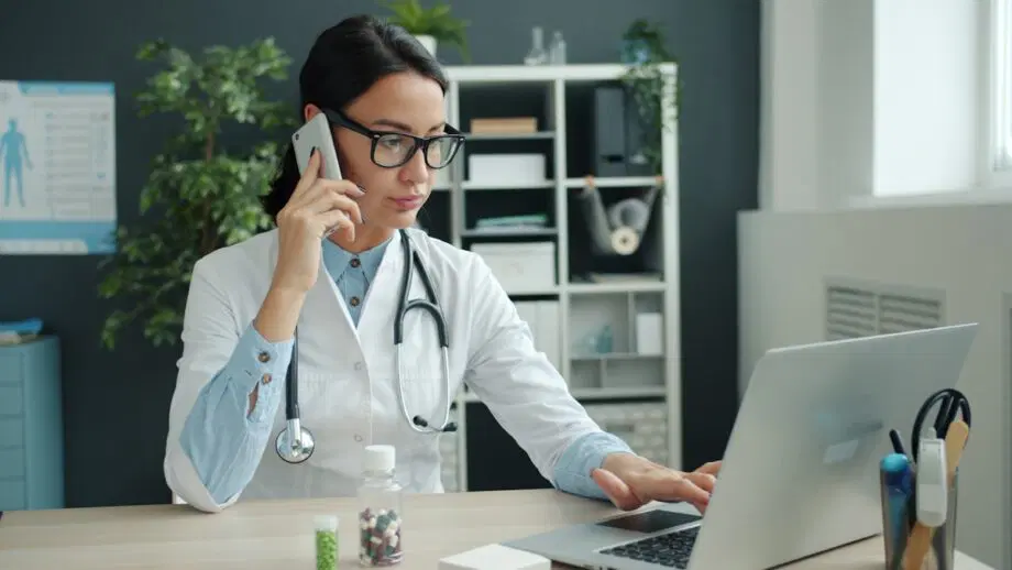 Un médecin en blouse blanche utilise un ordinateur portable et parle au téléphone à son bureau, avec des flacons de médicaments et des étagères de bureau en arrière-plan.