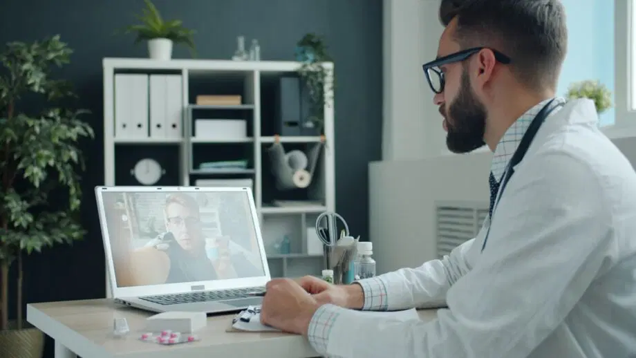 Un médecin en blouse blanche est assis à un bureau et mène un appel vidéo avec un patient sur un ordinateur portable dans un bureau moderne.