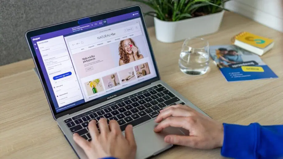 Personne utilisant un ordinateur portable avec une interface logicielle de conception à l’écran. Un verre d'eau, une plante et quelques brochures sont sur la table en bois.