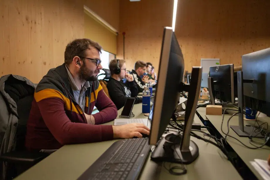 Plusieurs personnes portant des écouteurs travaillent d’affilée devant des ordinateurs de bureau à l’intérieur d’un bureau lambrissé ou d’un laboratoire informatique.