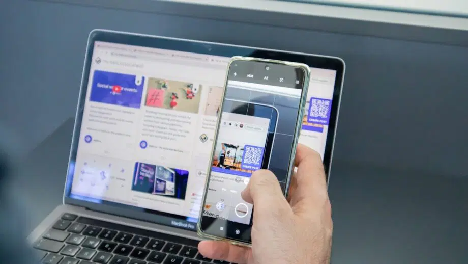 Personne tenant un smartphone pour scanner un code QR affiché sur l'écran d'un ordinateur portable affichant un site Web avec du contenu de médias sociaux.
