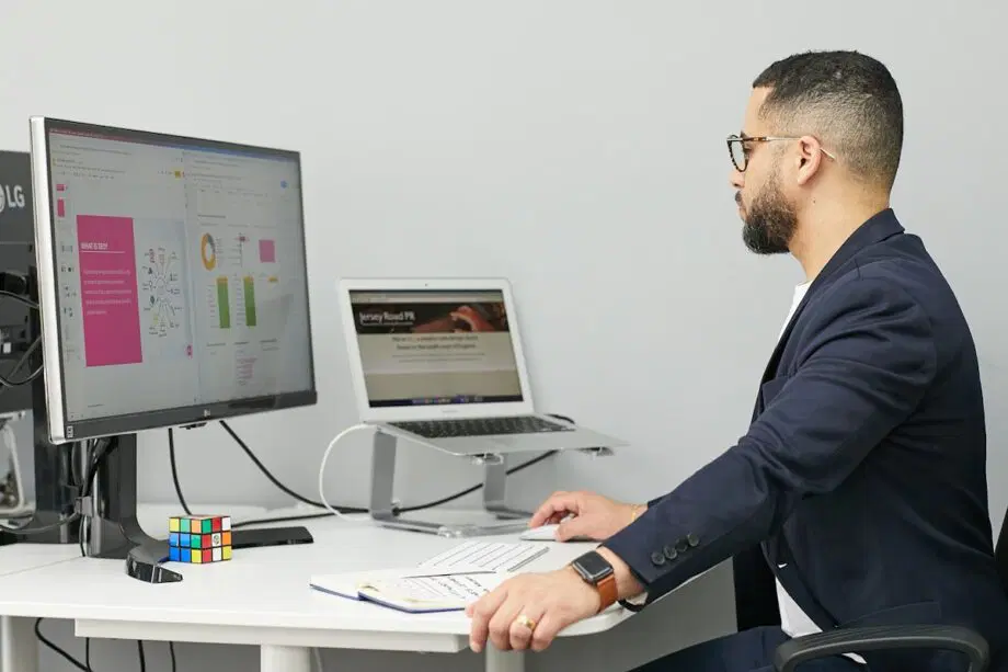 Un homme est assis à un bureau et travaille sur un ordinateur et un ordinateur portable, avec des documents, un cahier et un Rubik's Cube à proximité.