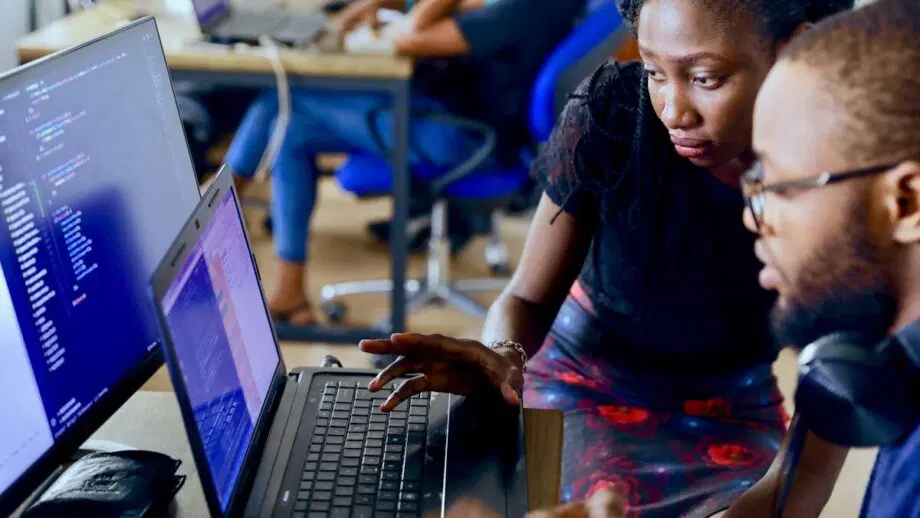 Deux personnes collaborent autour d'un bureau avec des ordinateurs portables, révisant le code à l'écran, tandis que d'autres travaillent en arrière-plan.