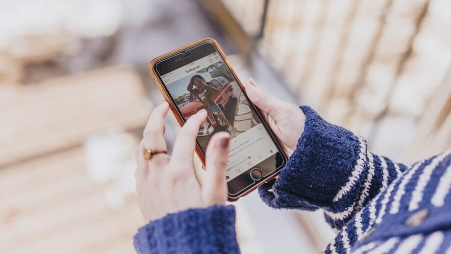 Personne vêtue d'un pull bleu tenant un smartphone et regardant une photo d'une femme sur Instagram à l'extérieur.