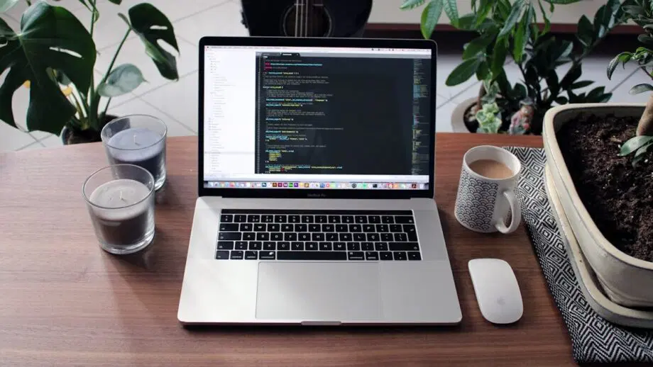 Un ordinateur portable affichant un code est posé sur une table en bois avec des bougies, une tasse, une souris d'ordinateur, des plantes d'intérieur et une guitare en arrière-plan.