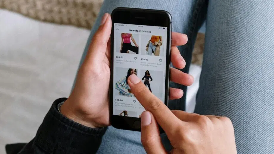 Une personne parcourt des vêtements sur un smartphone alors qu'elle est assise, les jambes croisées sur un lit.