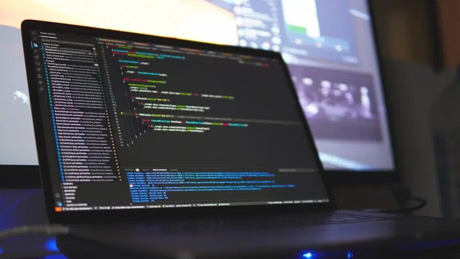 Un ordinateur portable affichant des lignes de code sur son écran, avec un autre moniteur flou en arrière-plan.