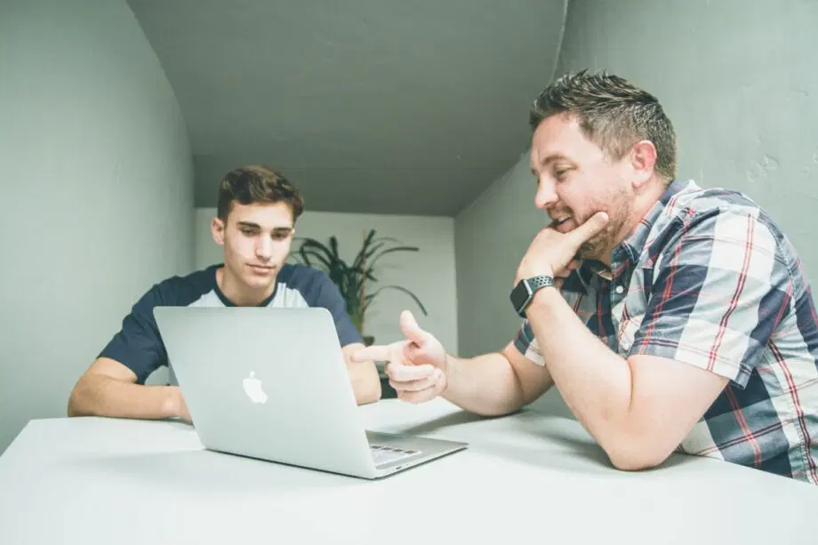 Deux hommes assis à une table regardent l’écran d’un ordinateur portable, l’un pointant vers l’écran pendant que l’autre écoute.