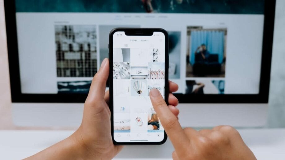 Gros plan d'une personne tenant un smartphone et parcourant une application de galerie d'images tandis qu'un écran d'ordinateur en arrière-plan affiche des images similaires.