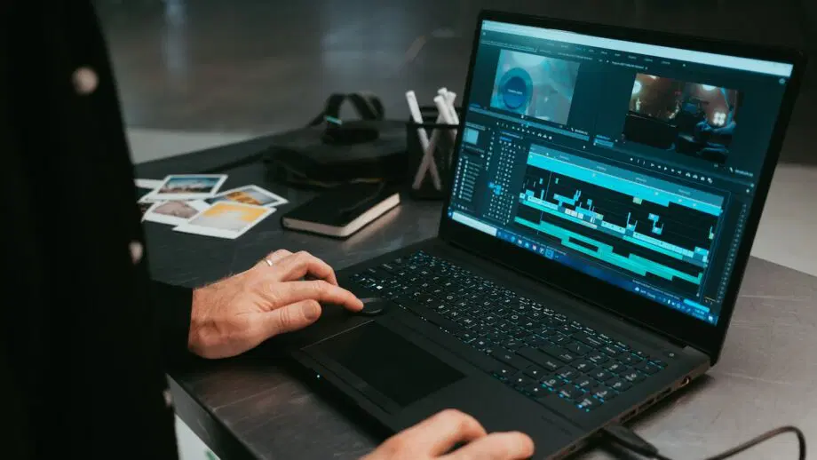 Personne utilisant un ordinateur portable avec un logiciel de montage vidéo ouvert, entourée de cahiers, de photos et d'accessoires électroniques sur un bureau.