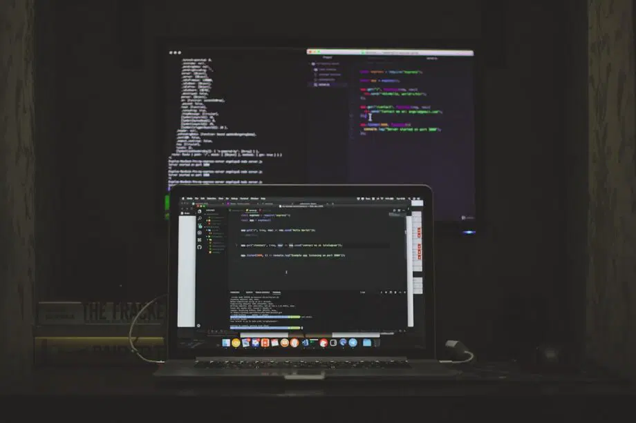 Un ordinateur portable et un moniteur affichent des lignes de code dans une pièce sombre, avec des livres empilés à côté de l'ordinateur portable.